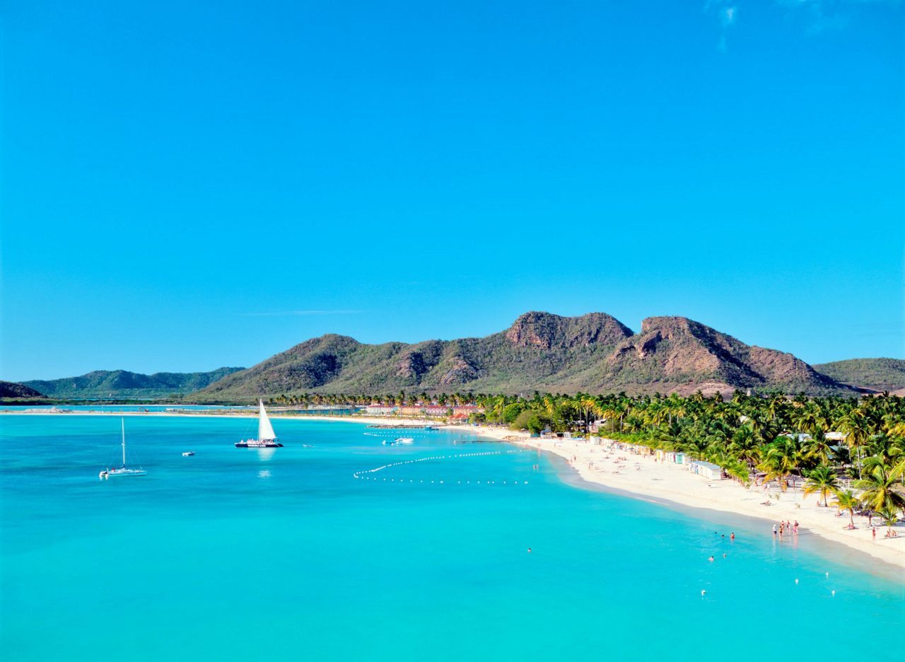 Aerial view of curved white sand beach with crystal clear turquoise water, palm trees, sailboats, and green hills under bright blue sky in Antigua.