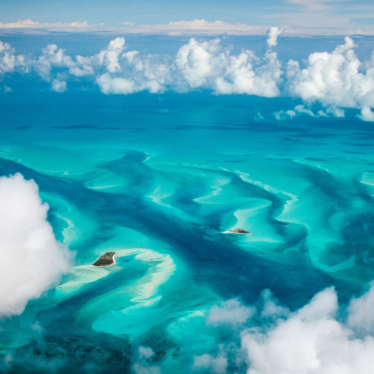 Aerial view of a coastal area at sunset with turquoise waters, scattered buildings, and lush greenery. Hills and islands are visible in the distance under a colorful sky.