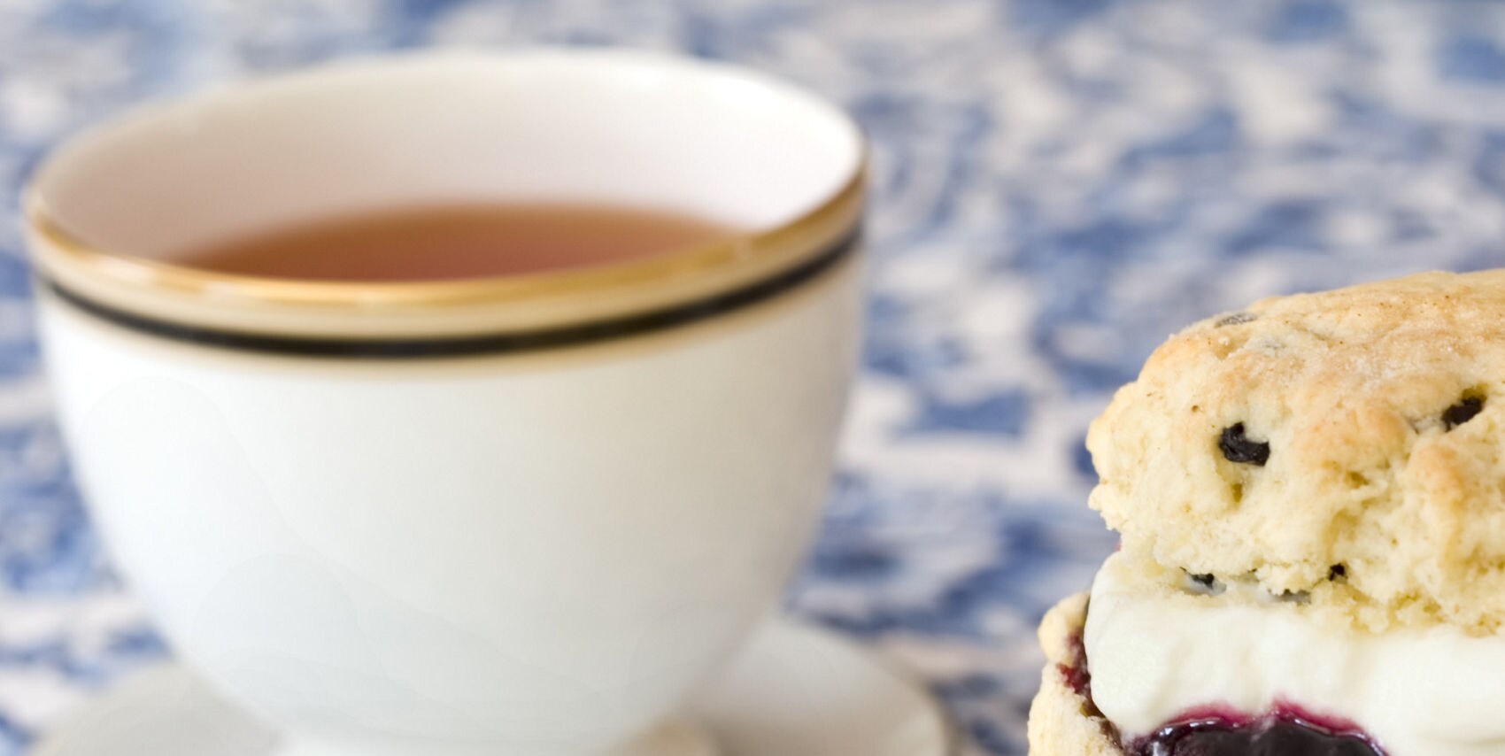 A cup of tea beside a stack of cream and jam-filled scones on a blue floral china plate.