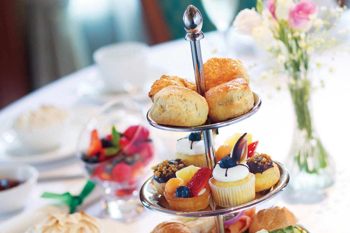 A tiered silver stand filled with scones, mini desserts, and fresh fruit displayed on a white-linen table.