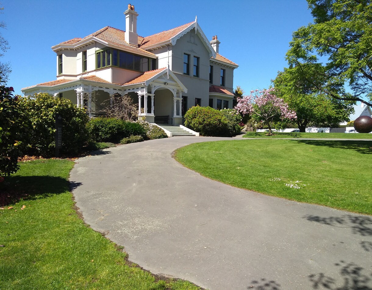 Aigantighe Art Gallery in Timaru, a large historic villa with a wraparound porch, surrounded by green lawns, flowering trees and bright spring foliage under a clear blue sky.