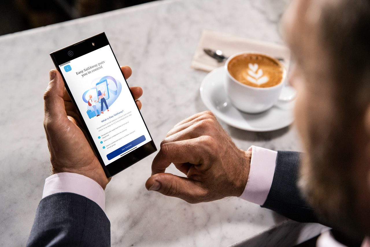 A close-up of a man's hands holding a smartphone displaying an app screen, with a latte on the table beside him.