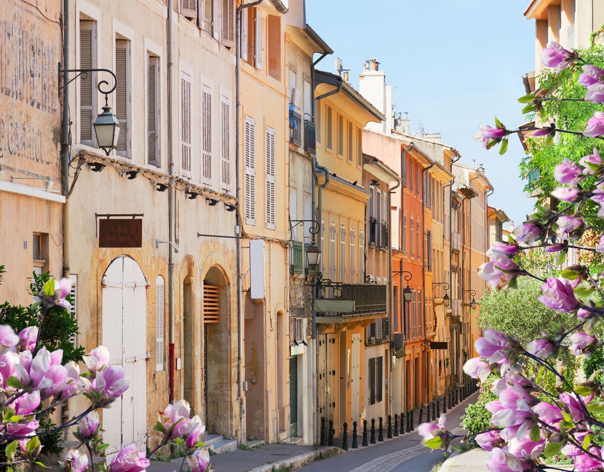 A narrow street in Aix-en-Provence with soft pastel buildings, shuttered windows and wrought-iron balconies, framed by pink spring blossoms in the foreground.