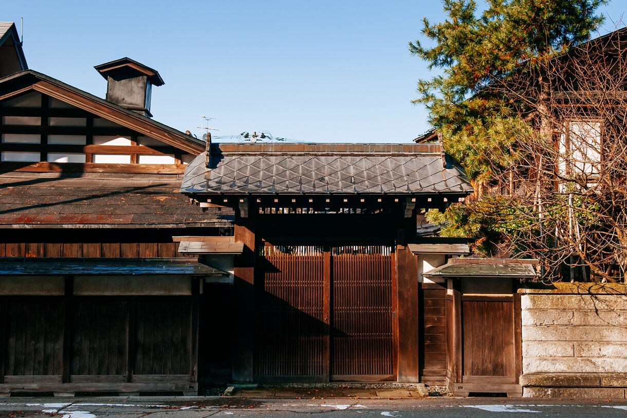 Traditional wooden gate and dark-tiled roof of a historic samurai house in Kakunodate, Japan, framed by trees and warm afternoon light.
