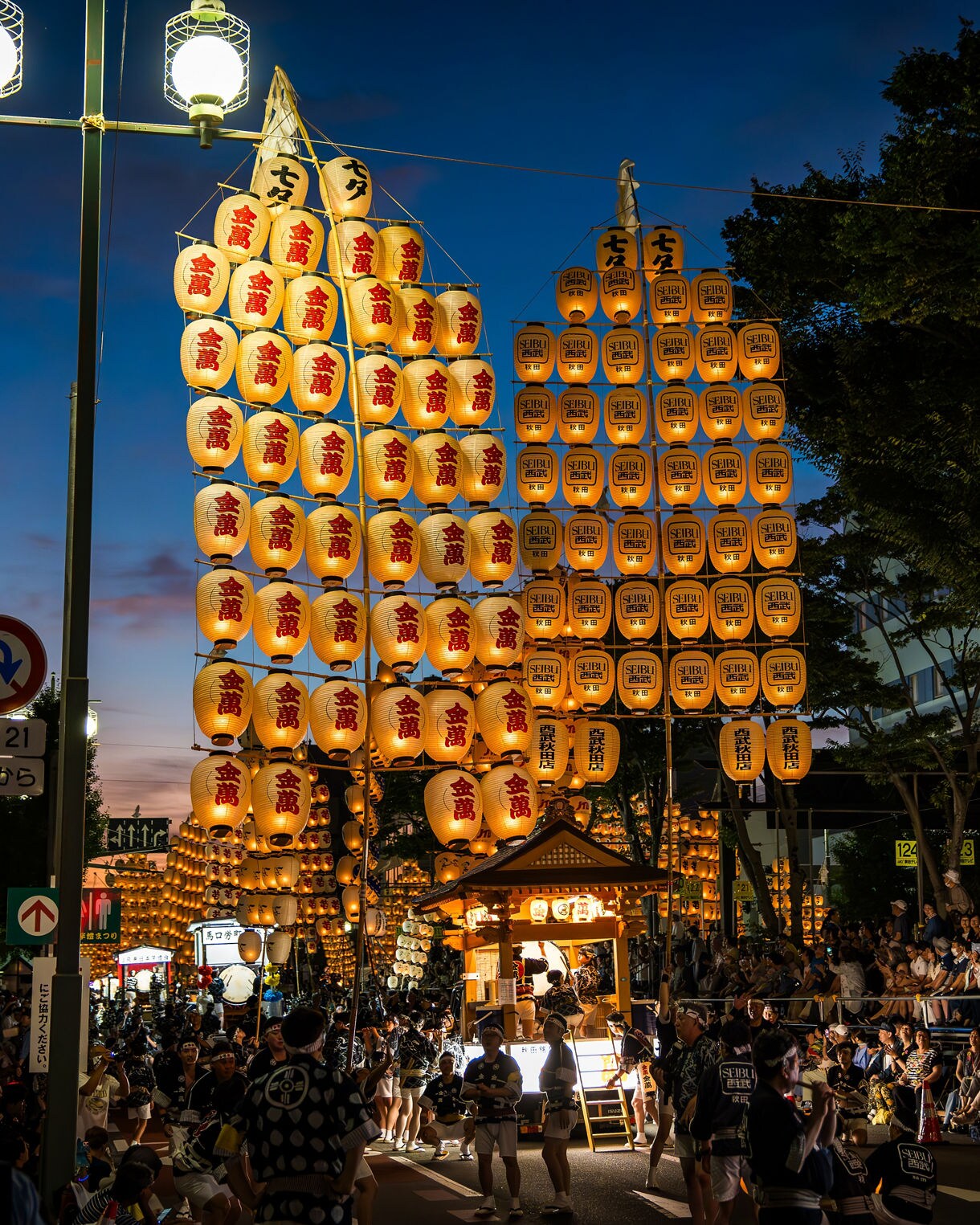 Nighttime street scene from Akita Kanto Festival with large poles of glowing lanterns balanced by performers and crowds watching from the sidelines.