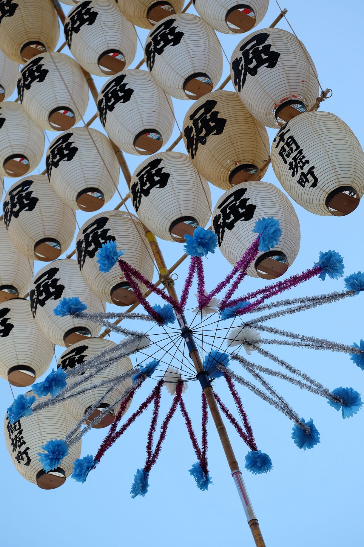 Looking up at numerous white paper lanterns with Japanese characters suspended around colorful tinsel streamers that extend radially from a central wooden pole, photographed from below against a bright blue sky.