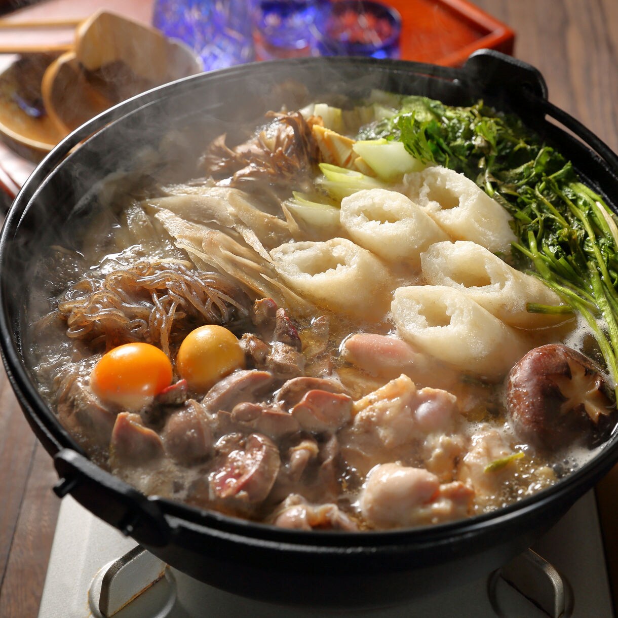 Close-up of kiritanpo nabe hot pot with rice skewers, chicken, mushrooms, greens, and vegetables simmering in broth.
