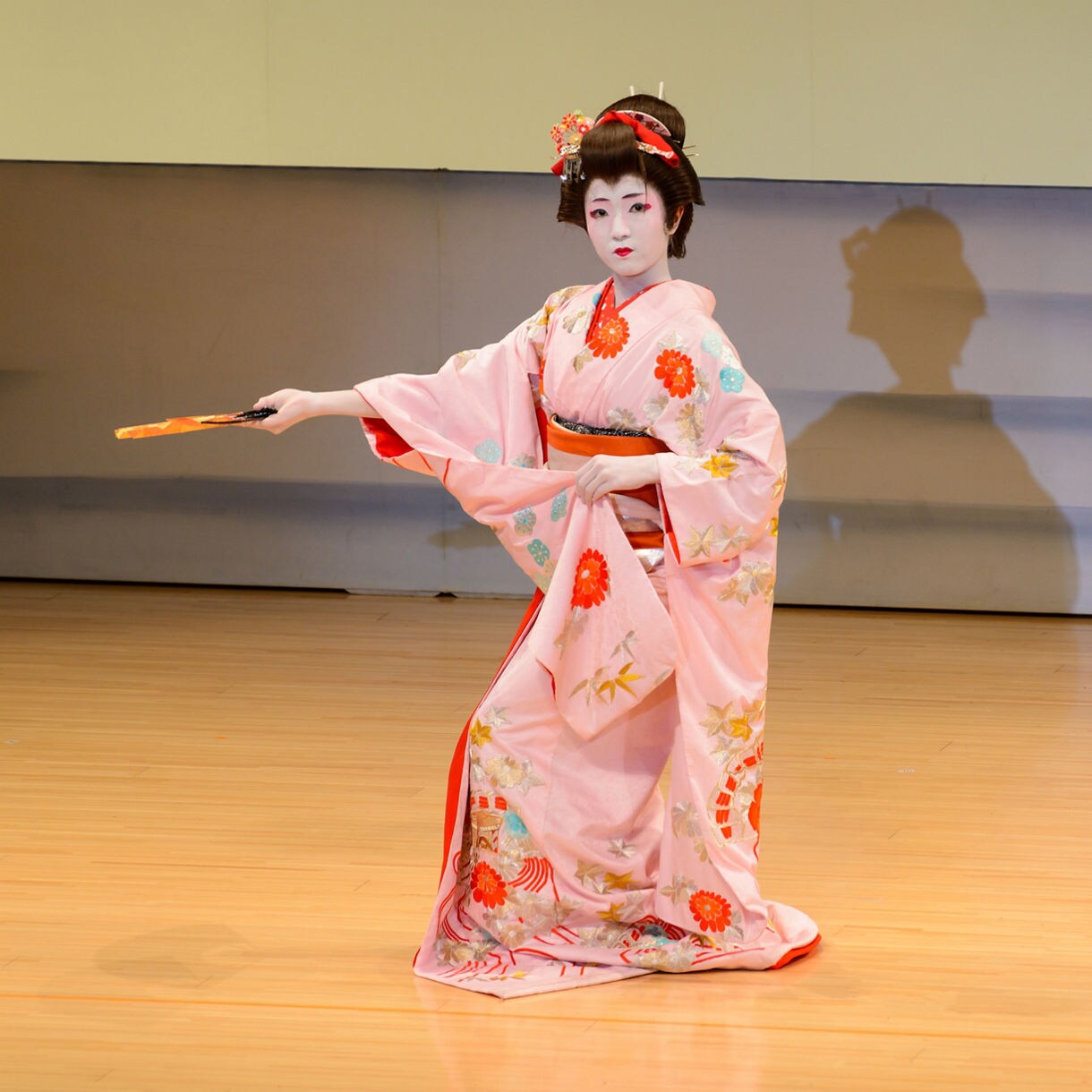 Maiko in a pink kimono performing a traditional Japanese dance with a fan on a wooden stage.
