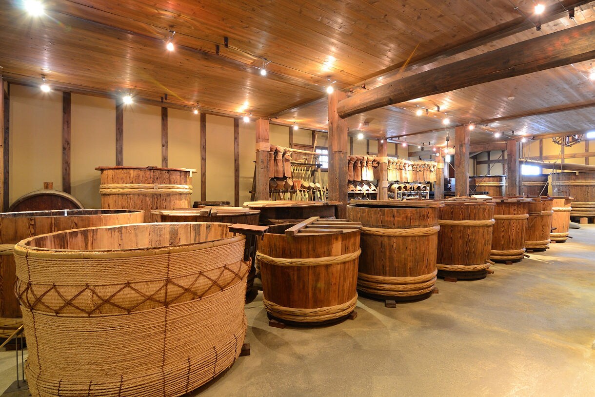 Interior of a traditional Japanese sake brewery with large wooden barrels and brewing tools lining the walls.