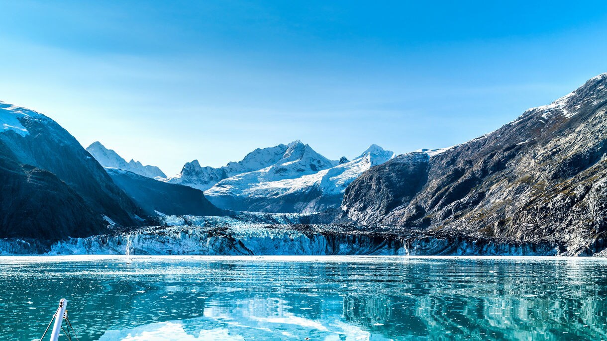 A scenic view of snow-capped mountains and a glacier reflected in calm blue waters with floating ice chunks in Alaska. 