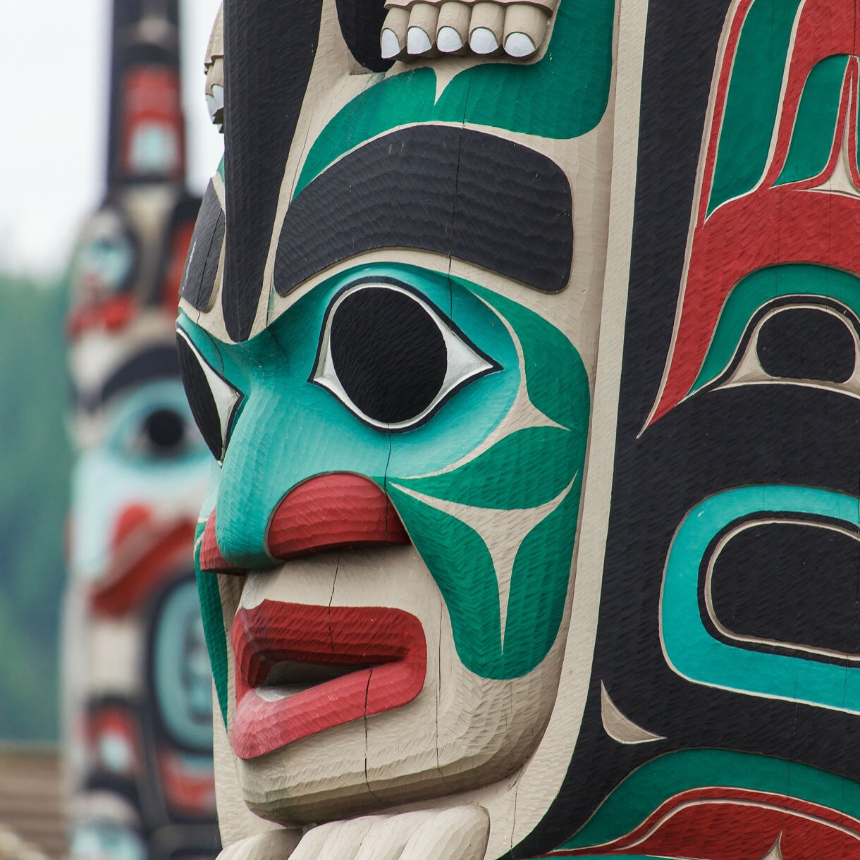 Dramatic close-up of a freshly carved and painted Alaska Native totem pole in teal, red and black with additional totem poles and a forested hillside blurred in the background.