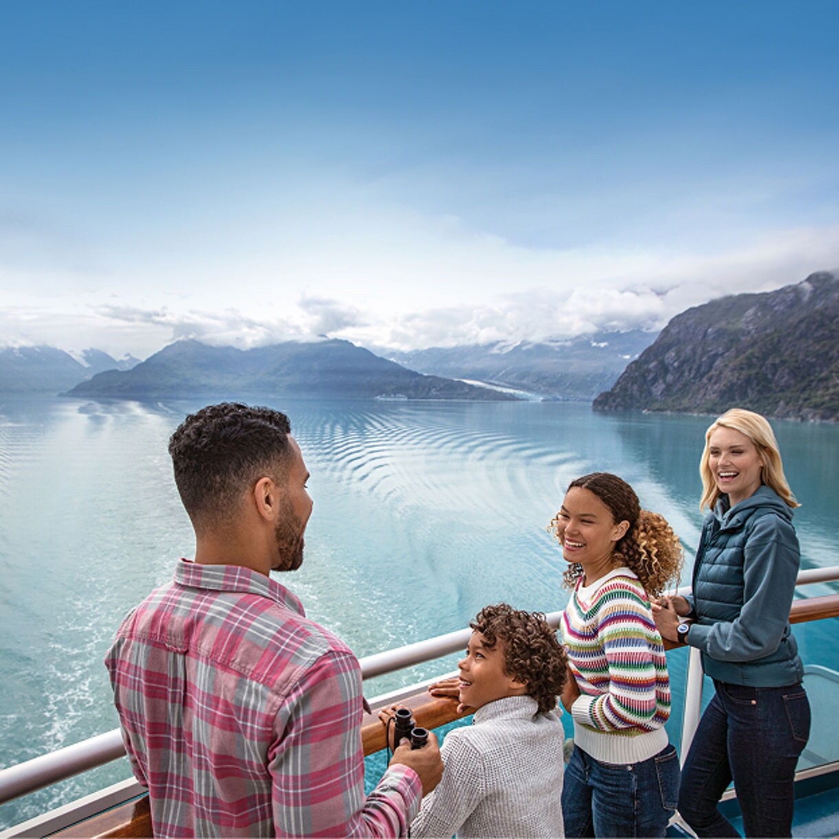 A group of people stands on the deck of a cruise ship, gazing out over a calm, expansive body of water surrounded by mountains. The setting is outdoors with a serene, natural landscape and clear blue sky. The group includes adults and children, dressed casually, suggesting a relaxed vacation atmosphere. The overall mood is peaceful and joyful, with cool tones dominating the scene.