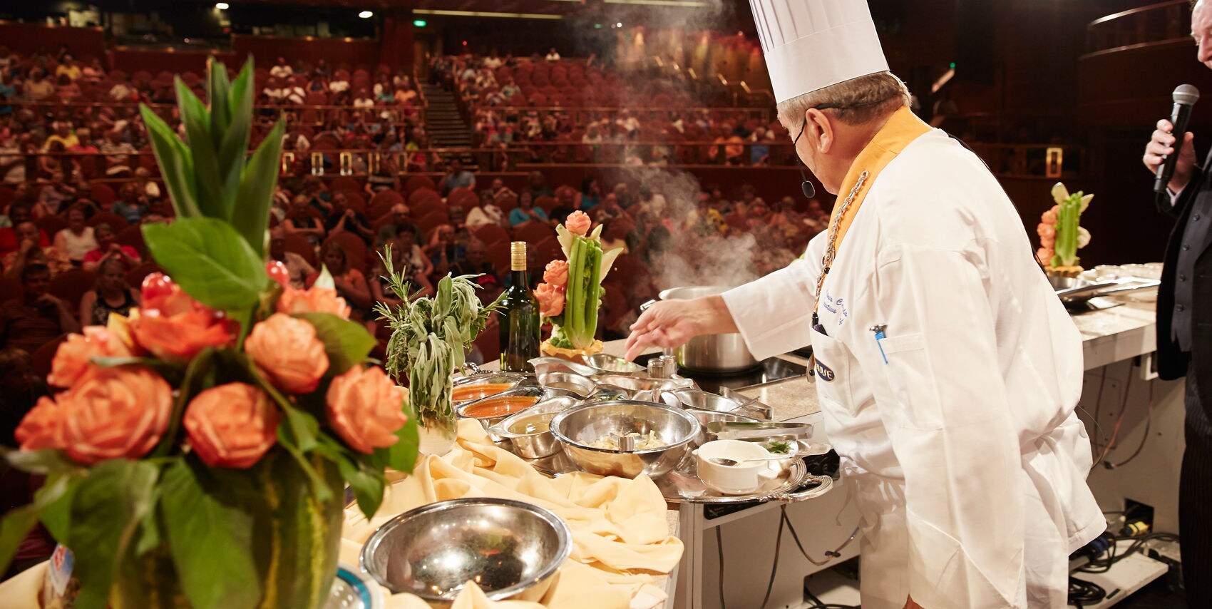 A professional chef in a white uniform and tall hat prepares food on a stage during a live cooking demonstration. The setting is a large auditorium filled with an audience, with bright stage lights illuminating the scene. Fresh ingredients and floral arrangements are visible on the table, and steam rises from the cooking pots. The atmosphere is lively and engaging, highlighting culinary expertise.