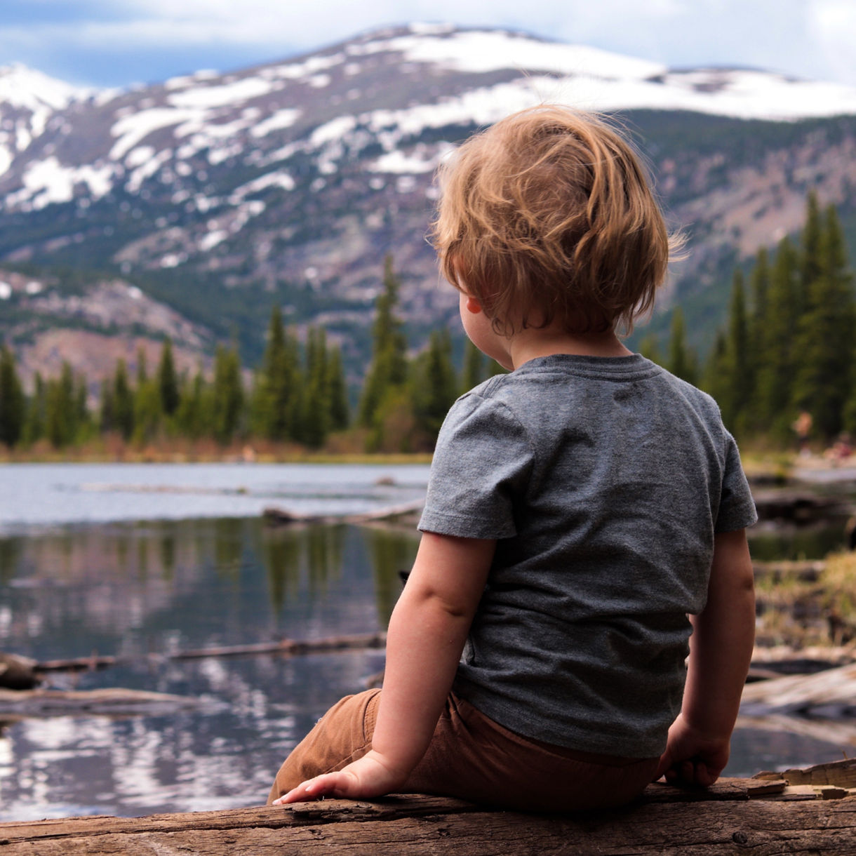 A small child sits on a log facing a calm lake surrounded by evergreen trees. Snow-capped mountains rise in the background under a partly cloudy sky. The child wears a gray t-shirt and brown pants, gazing toward the scenic wilderness. The scene conveys tranquility, exploration, and connection with nature.