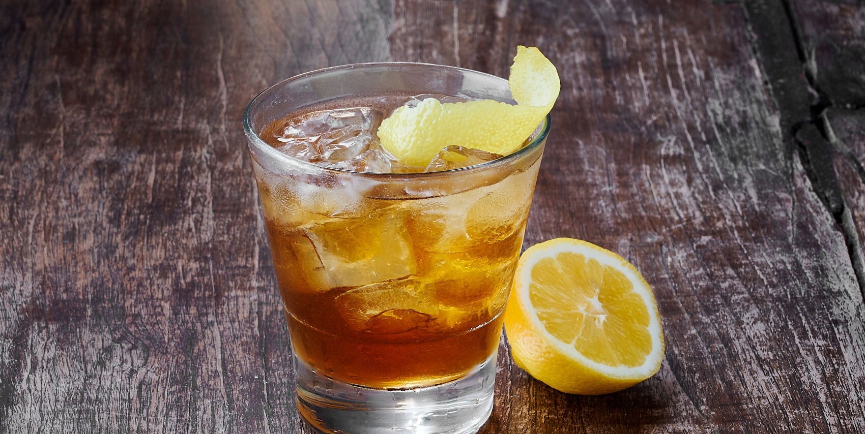 A rocks glass filled with an amber cocktail and ice, garnished with a lemon twist, beside a halved lemon on a weathered wood surface.