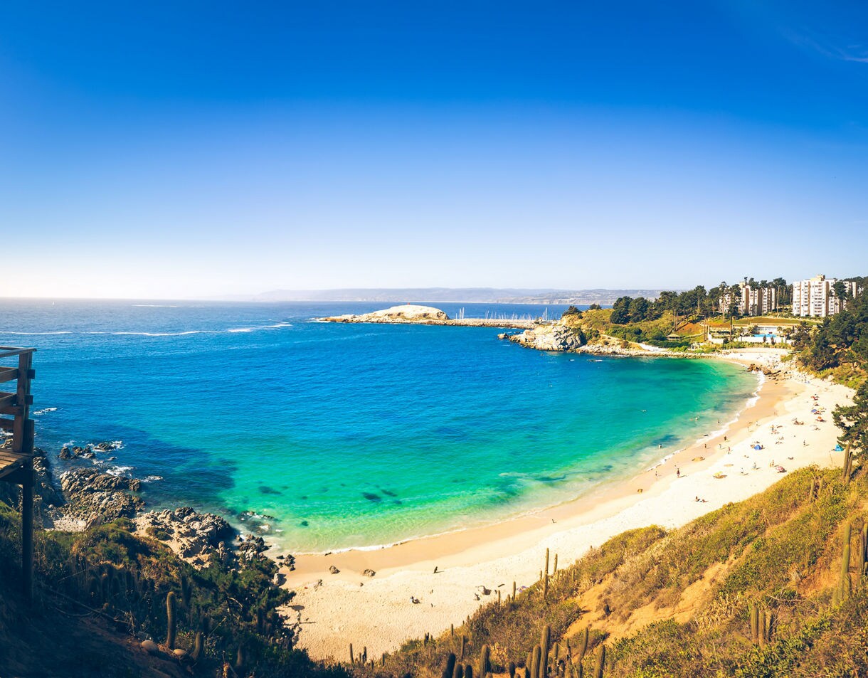 Panoramic view of a bright turquoise bay with a sandy beach, rocky shoreline and buildings set against a clear blue sky.