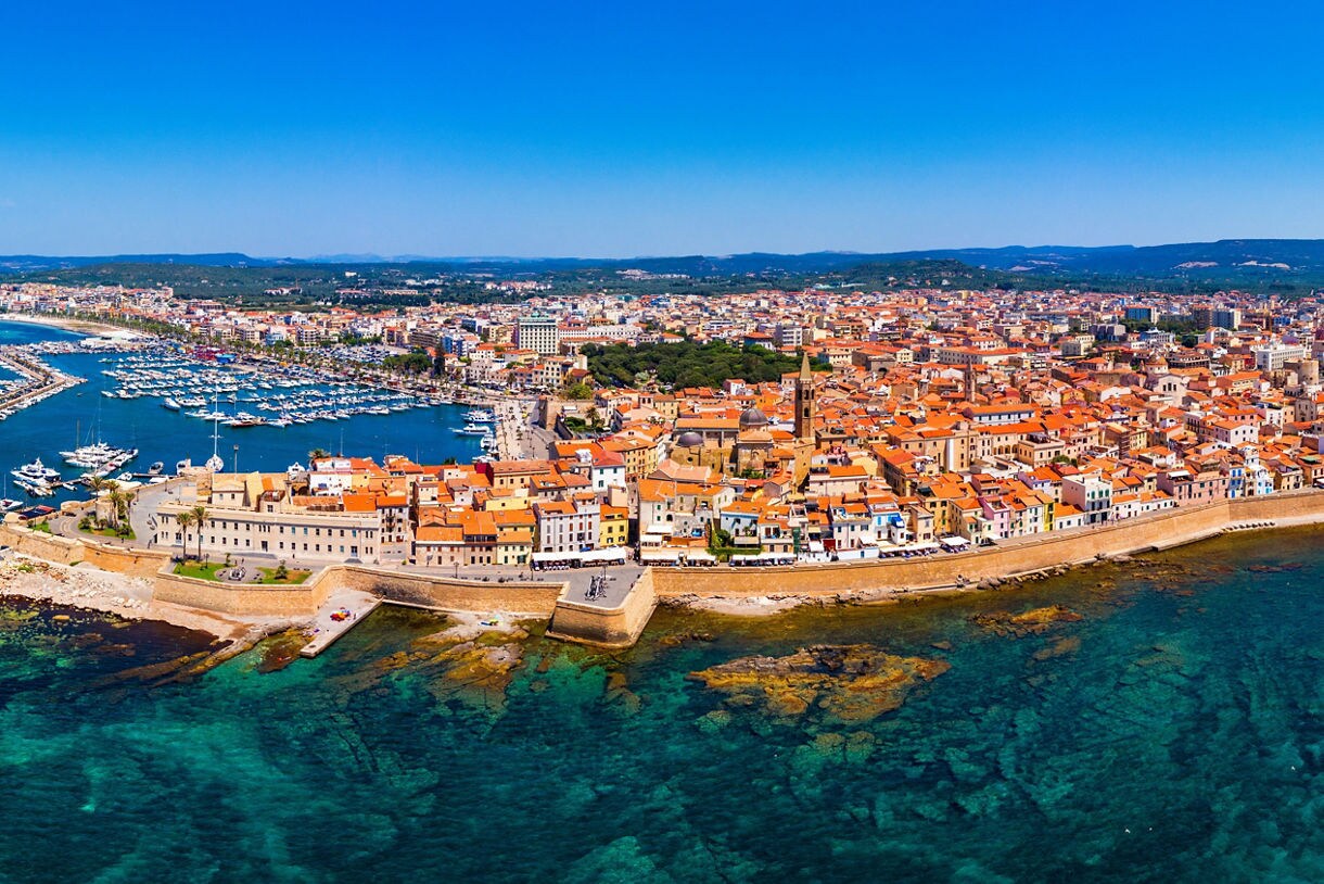 Aerial view of a coastal town with orange rooftops, stone sea walls and clear turquoise water.
