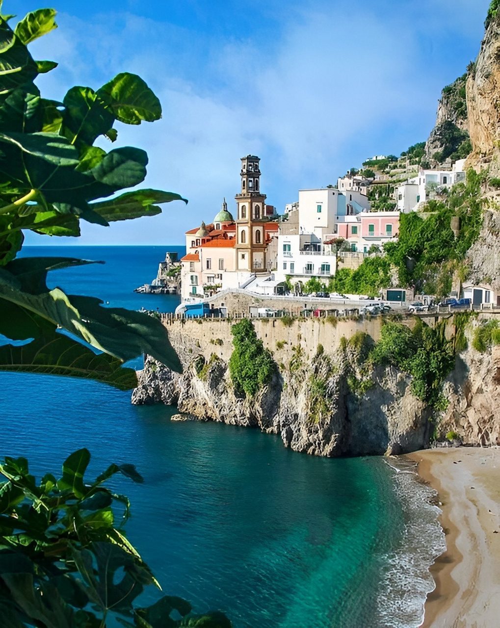A coastal village on the Amalfi Coast with pastel buildings perched on rocky cliffs above a small sandy beach and vivid blue water, framed by green leaves.