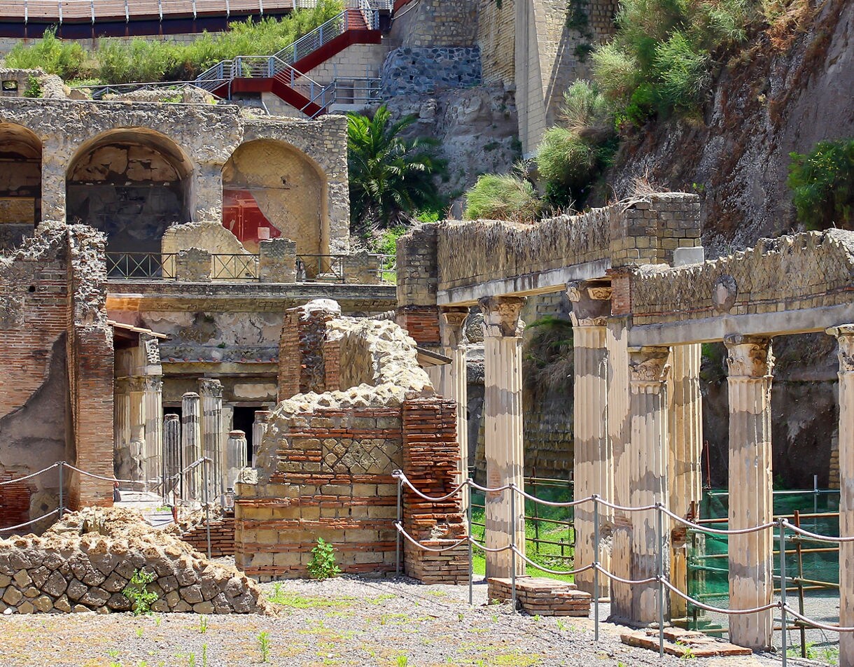 Ruins of the ancient Roman town Herculaneum, showing weathered marble columns and brick structures excavated from the volcanic rock at the base of a cliffside.
