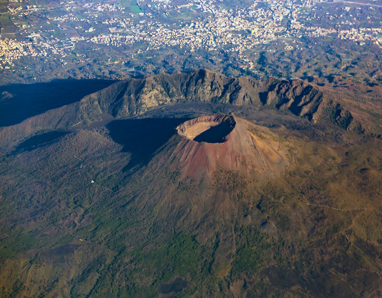 High aerial view directly over the volcanic cone of Mount Vesuvius, showing the large crater rim and the densely populated city sprawling across the surrounding landscape.