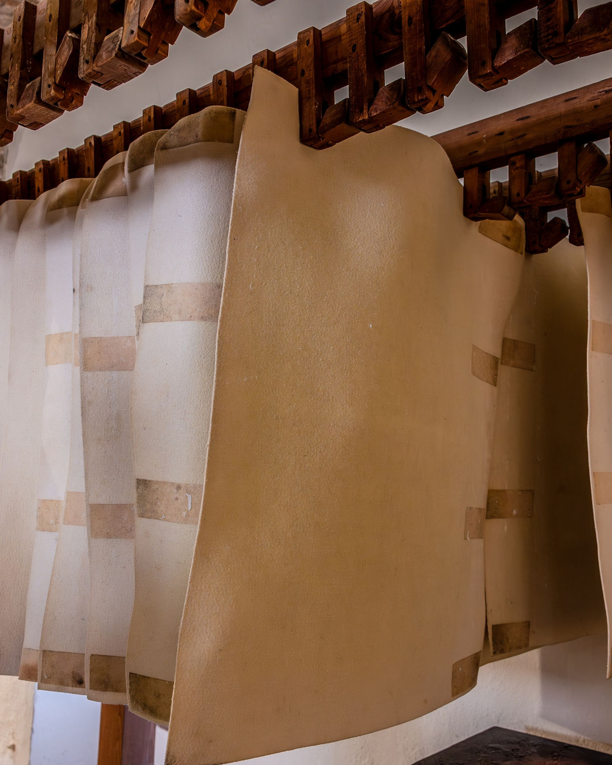 Sheets of handmade paper hang from thick wooden beams to dry inside a historic paper mill, with one sheet appearing slightly darker and closer to the foreground.