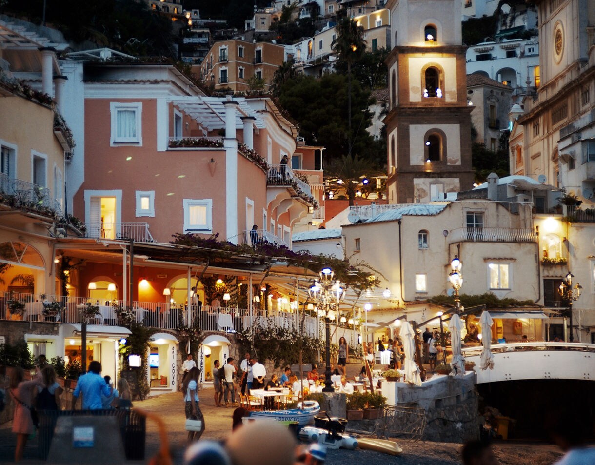 The seaside village of Positano at dusk, showing multi-story pastel buildings with illuminated windows and outdoor restaurants crowded with people near the water's edge.