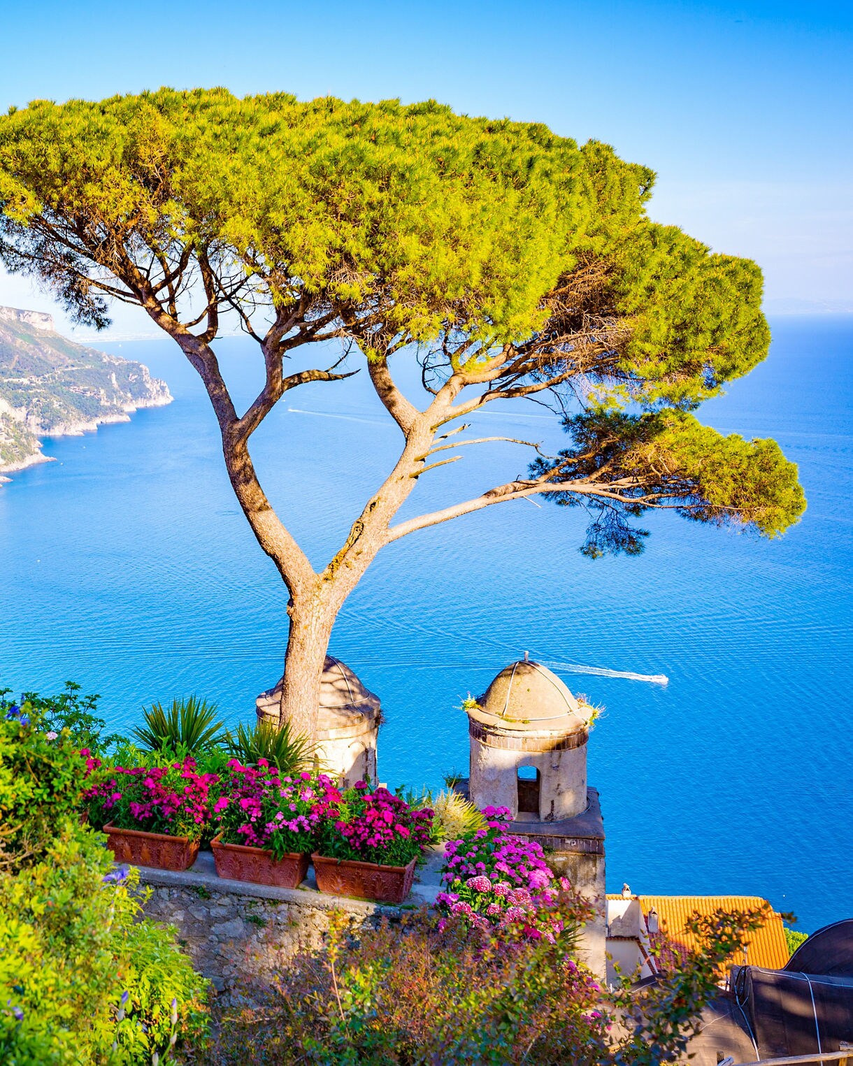 A panoramic view from the gardens of a villa in Ravello, showing a lone umbrella pine tree and a small stone turret overlooking the brilliant blue Tyrrhenian Sea and the rugged Amalfi Coastline.