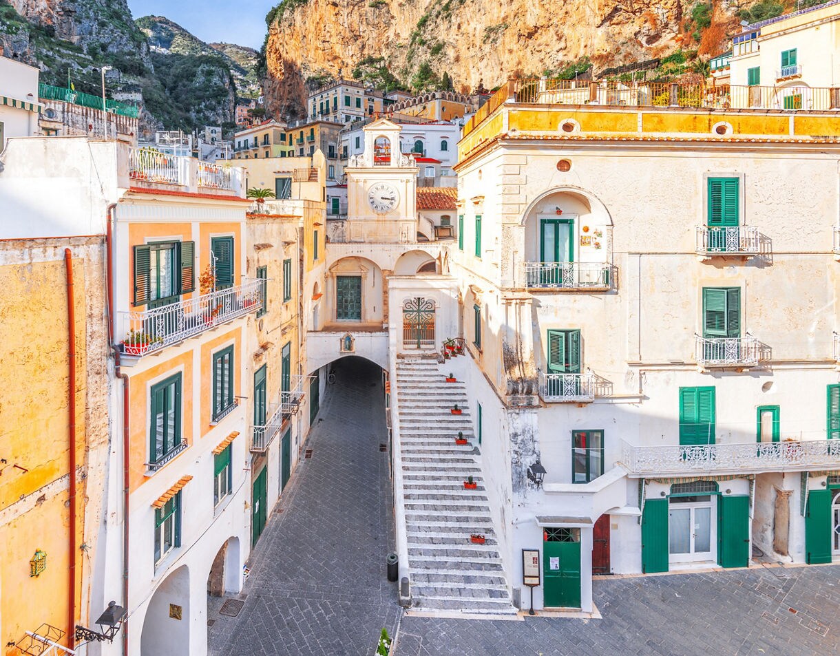 A narrow cobblestone alleyway in the historic center of an Italian town, flanked by tall buildings with green shutters, leading to a small bell tower and a clock face above a set of stairs.