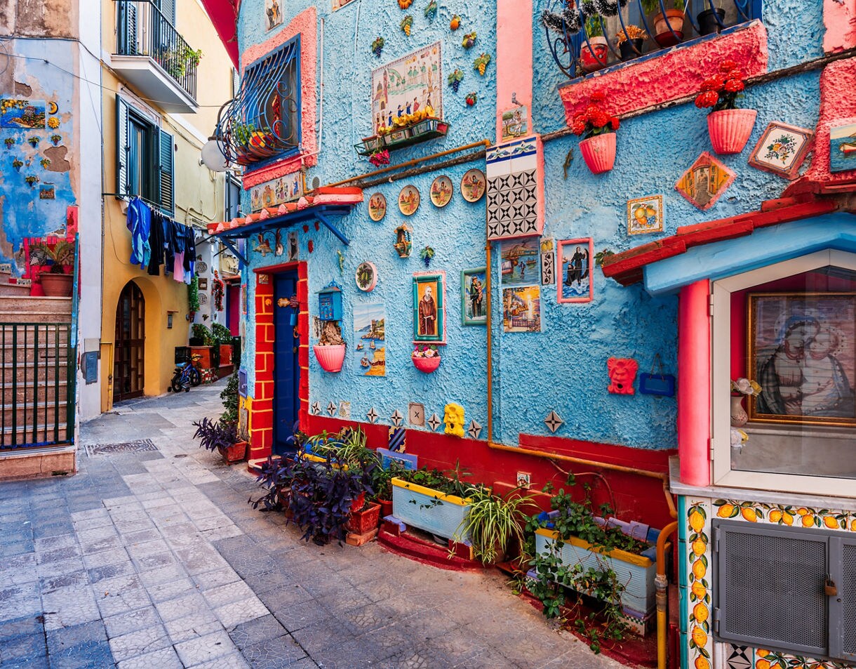 A narrow cobblestone alley in Vietri sul Mare, Italy, featuring a building facade painted bright blue and decorated with numerous colorful ceramic tiles, terracotta pots, and pink trim.