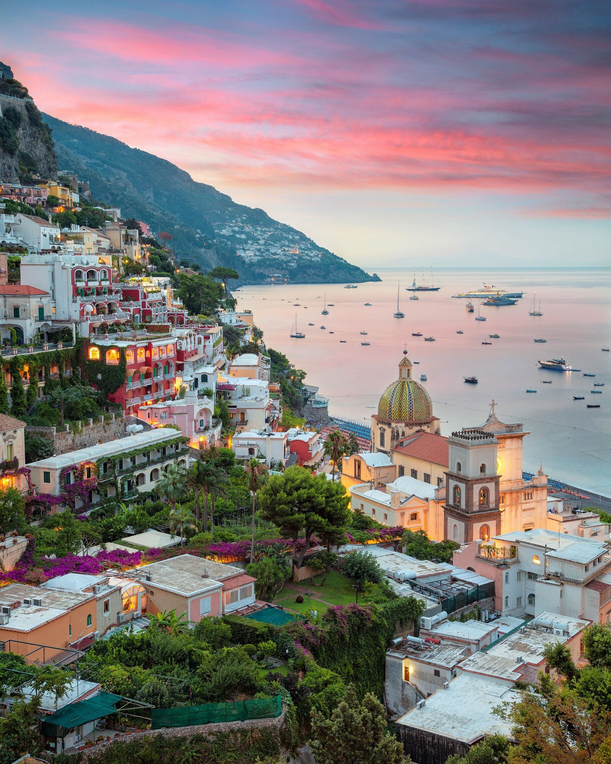 High-angle vertical view of the village of Positano at twilight, with lights illuminating the houses and the dome of the Church of Santa Maria Assunta, facing the sea dotted with numerous anchored boats under a sky streaked with pink and purple clouds.