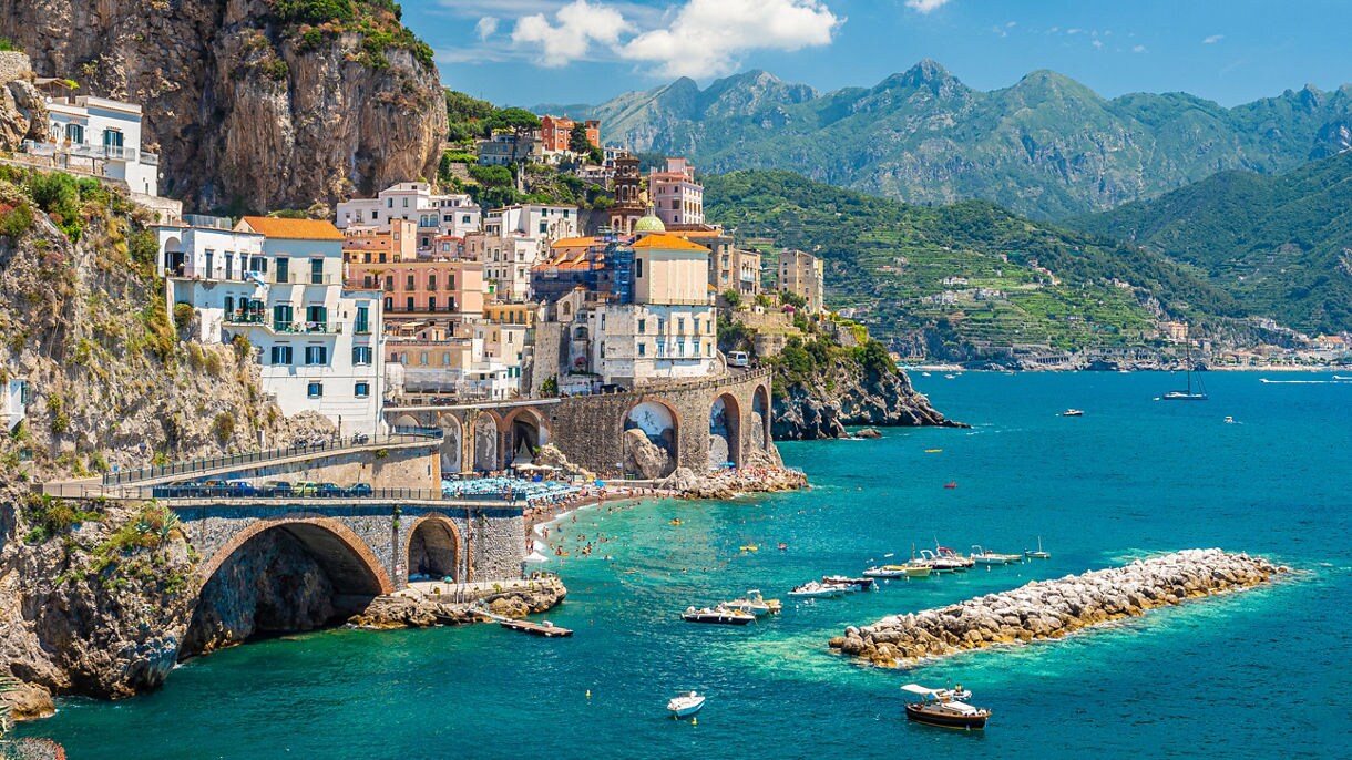 A stunning view of the Amalfi Coast in Italy, showcasing colorful buildings perched on cliffs overlooking the turquoise sea, with small boats docked along the shoreline and mountains in the background.