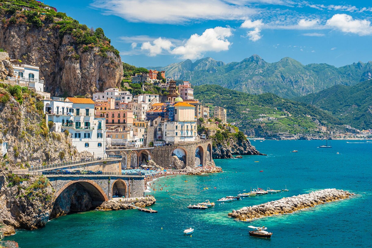 A stunning view of the Amalfi Coast in Italy, showcasing colorful buildings perched on cliffs overlooking the turquoise sea, with small boats docked along the shoreline and mountains in the background.