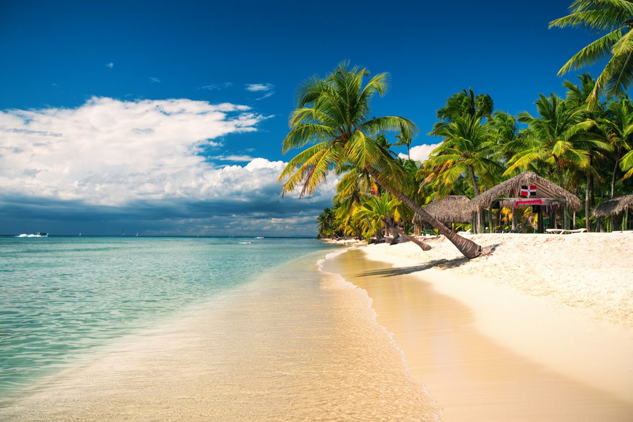 A serene beach in Amber Cove, Dominican Republic featuring clear shallow water, white sand, leaning palm trees and a thatched-roof hut with a Dominican flag.