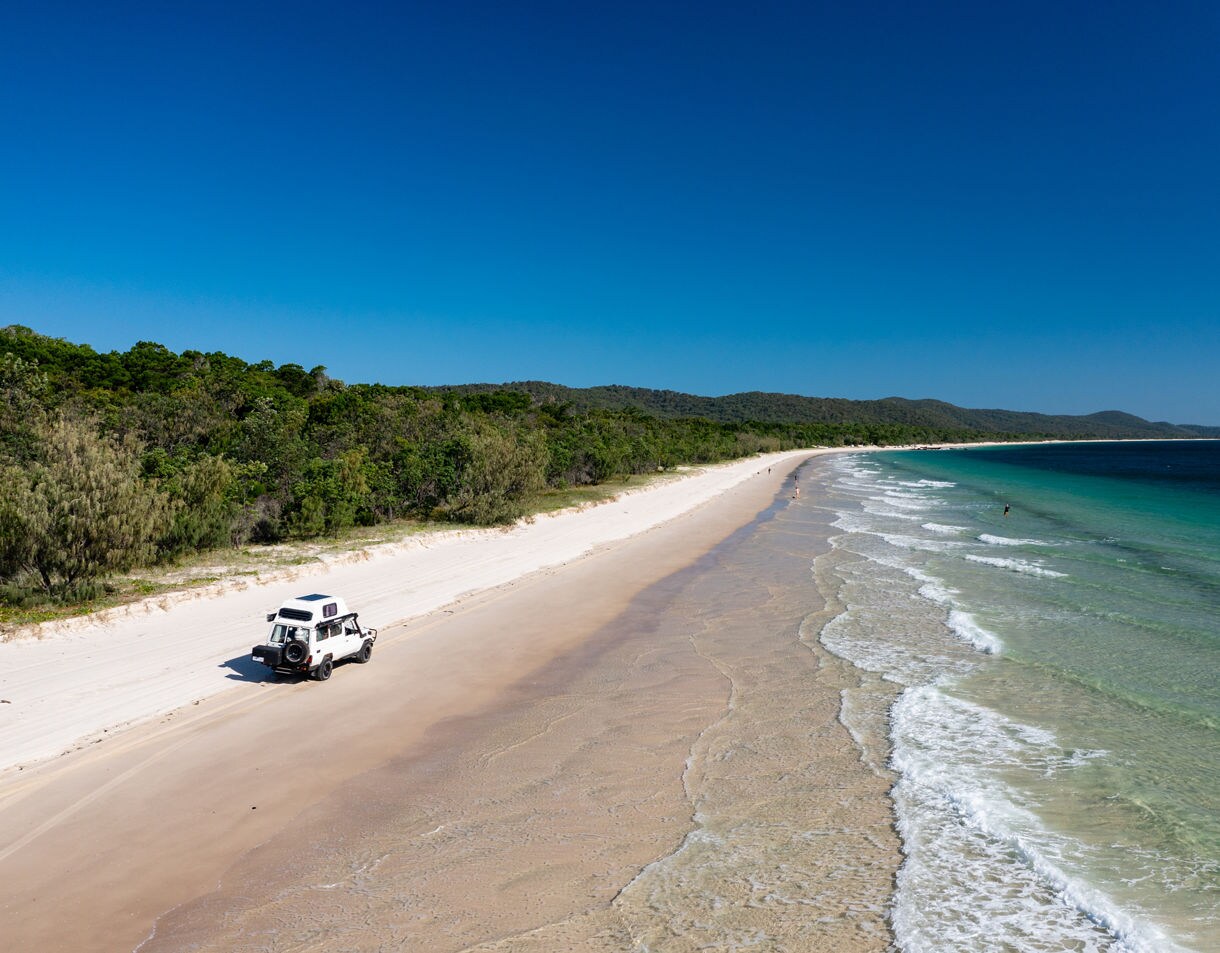 A white off-road truck driving on a wide sandy beach lined with lush green forest.