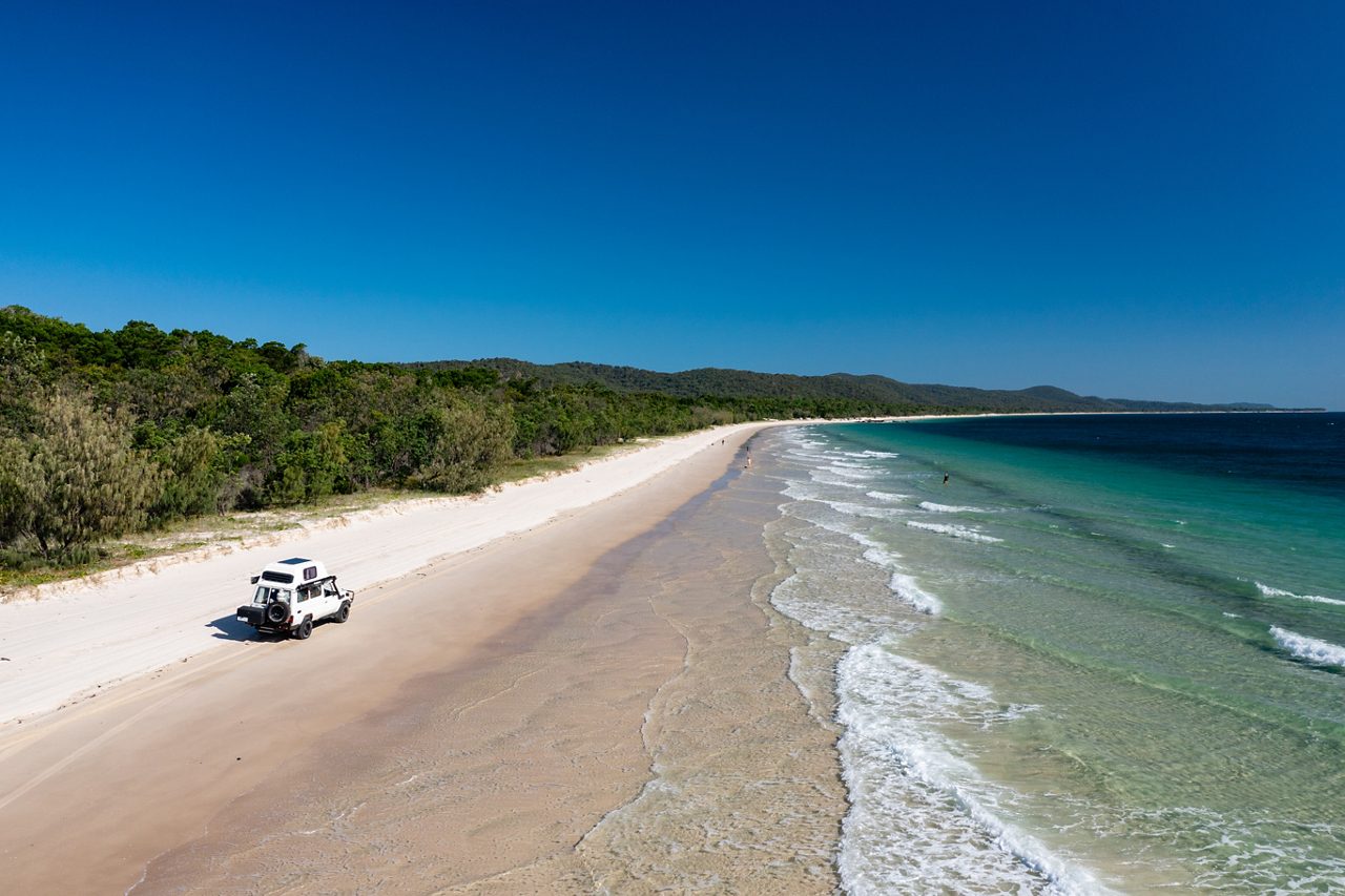 A white off-road truck driving on a wide sandy beach lined with lush green forest.