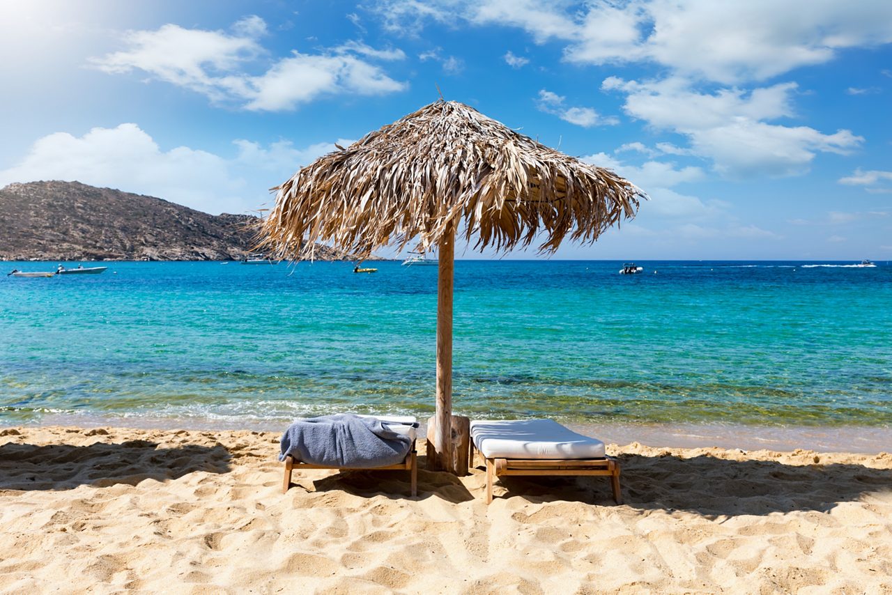 Two lounge chairs under a thatched umbrella on a sandy beach facing the clear blue waters of Amber Cove, with boats in the distance and a rocky hillside across the bay.