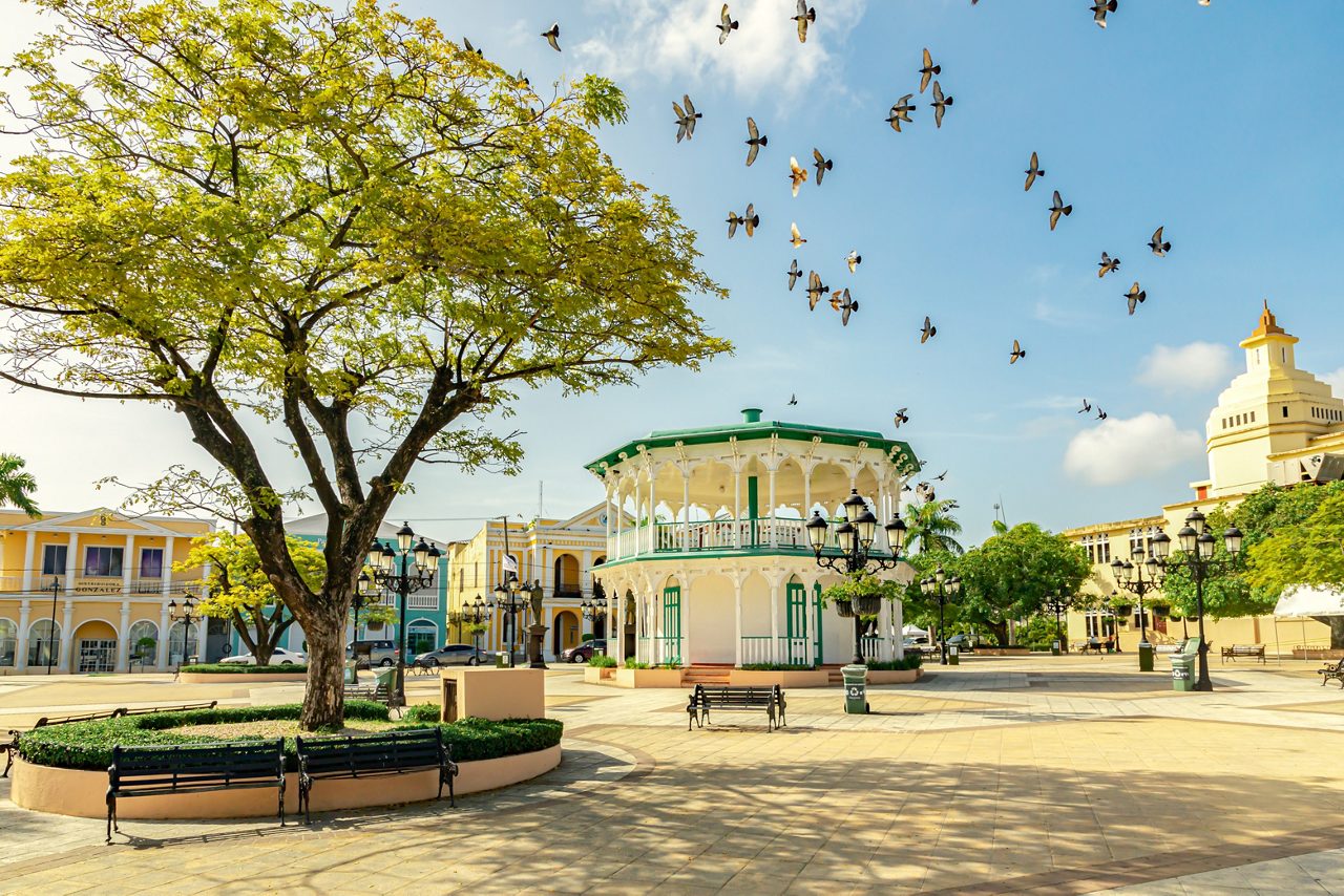 A scenic plaza in Puerto Plata, Dominican Republic featuring a white and green Victorian-style gazebo, a large tree with yellow-green leaves, colonial buildings and pigeons soaring against a bright blue sky.