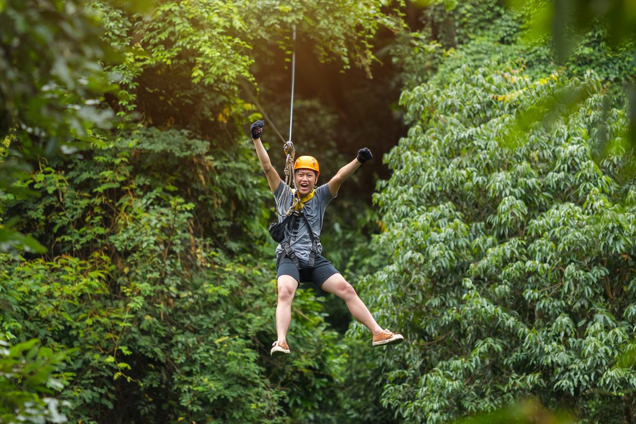 Person ziplining through a lush tropical forest in Amber Cove, Dominican Republic, wearing a helmet and safety harness with arms raised in excitement.