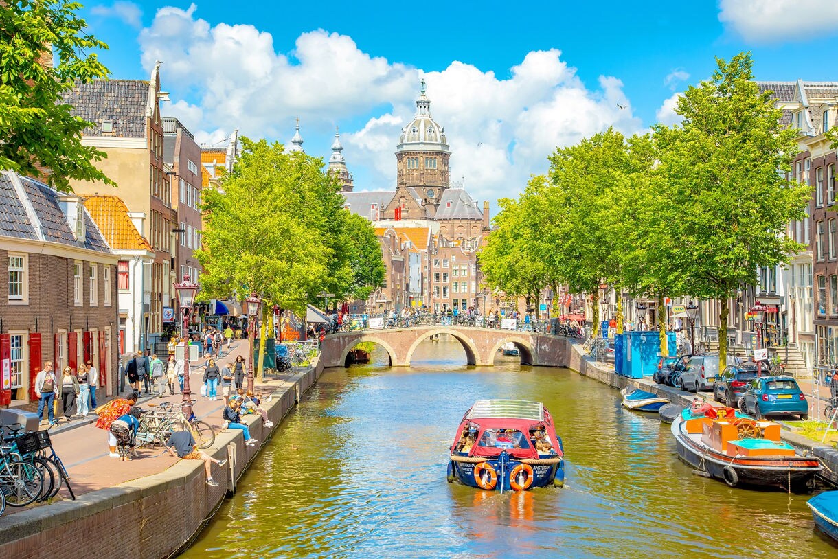 Canal boat passing under a stone bridge in central Amsterdam with historic buildings, trees and a church dome.