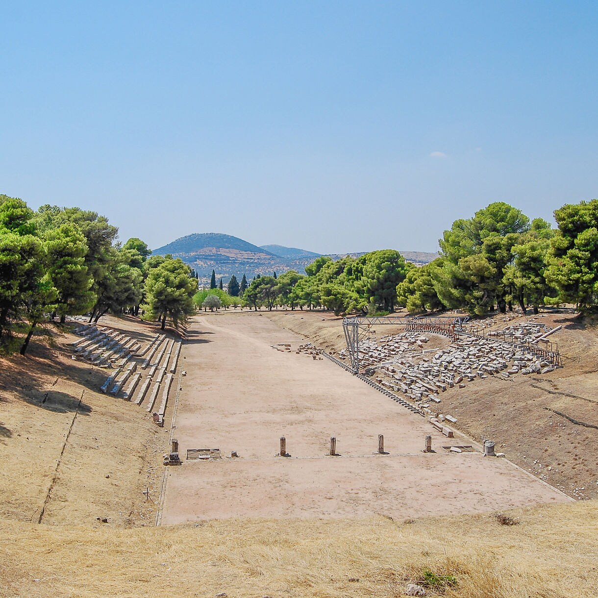 Panoramic view of the ancient Olympic stadium in Greece, with stone seating, dry grassy hills and rows of trees leading toward distant mountains under a clear blue sky.