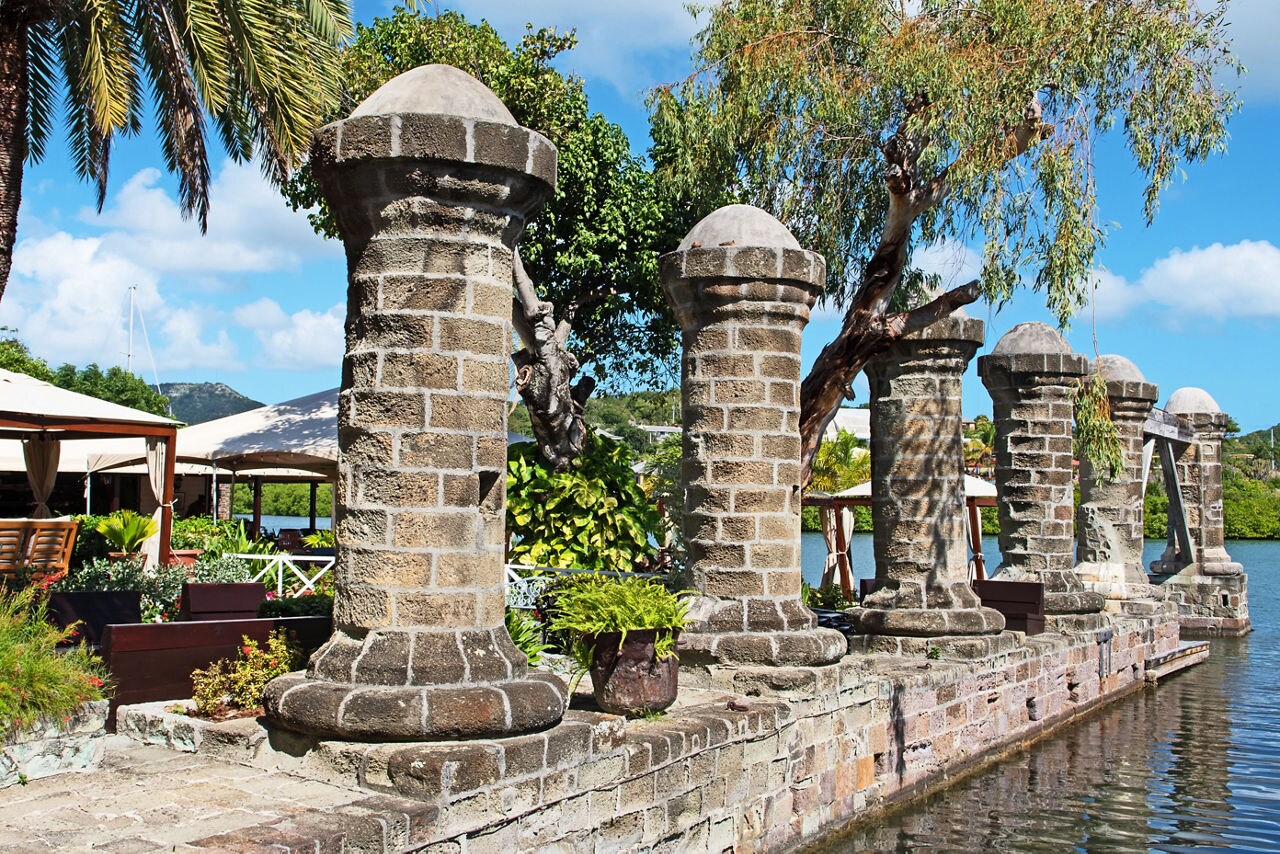 Historic stone pillars at Nelson’s Dockyard in Antigua standing along the waterfront with lush greenery, outdoor seating and sailboats in the background under a bright blue sky.