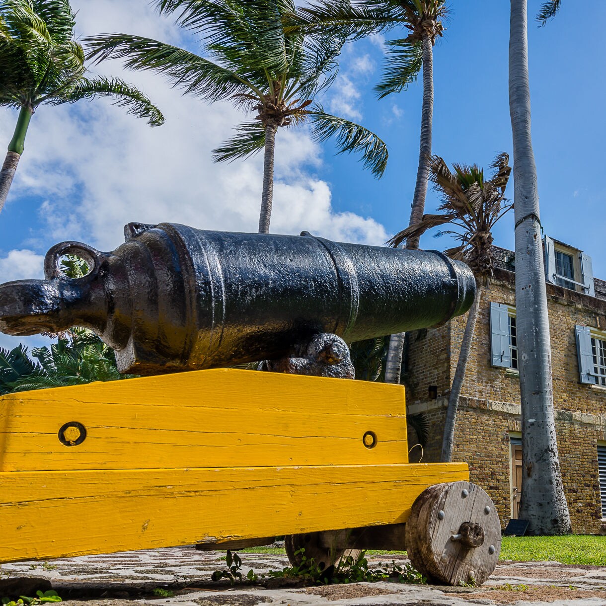 Historic black cannon on a bright yellow base in front of a stone colonial-era building with open shutters and tall palm trees at Nelson’s Dockyard in Antigua.
