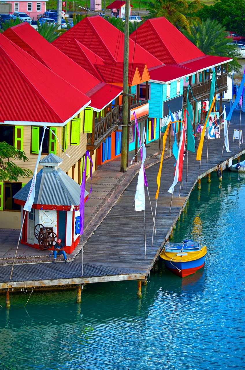 A row of brightly colored buildings with red roofs lining a wooden dock in St. John's, Antigua. Colorful flags hang along the dock next to a small boat moored in the turquoise water.
