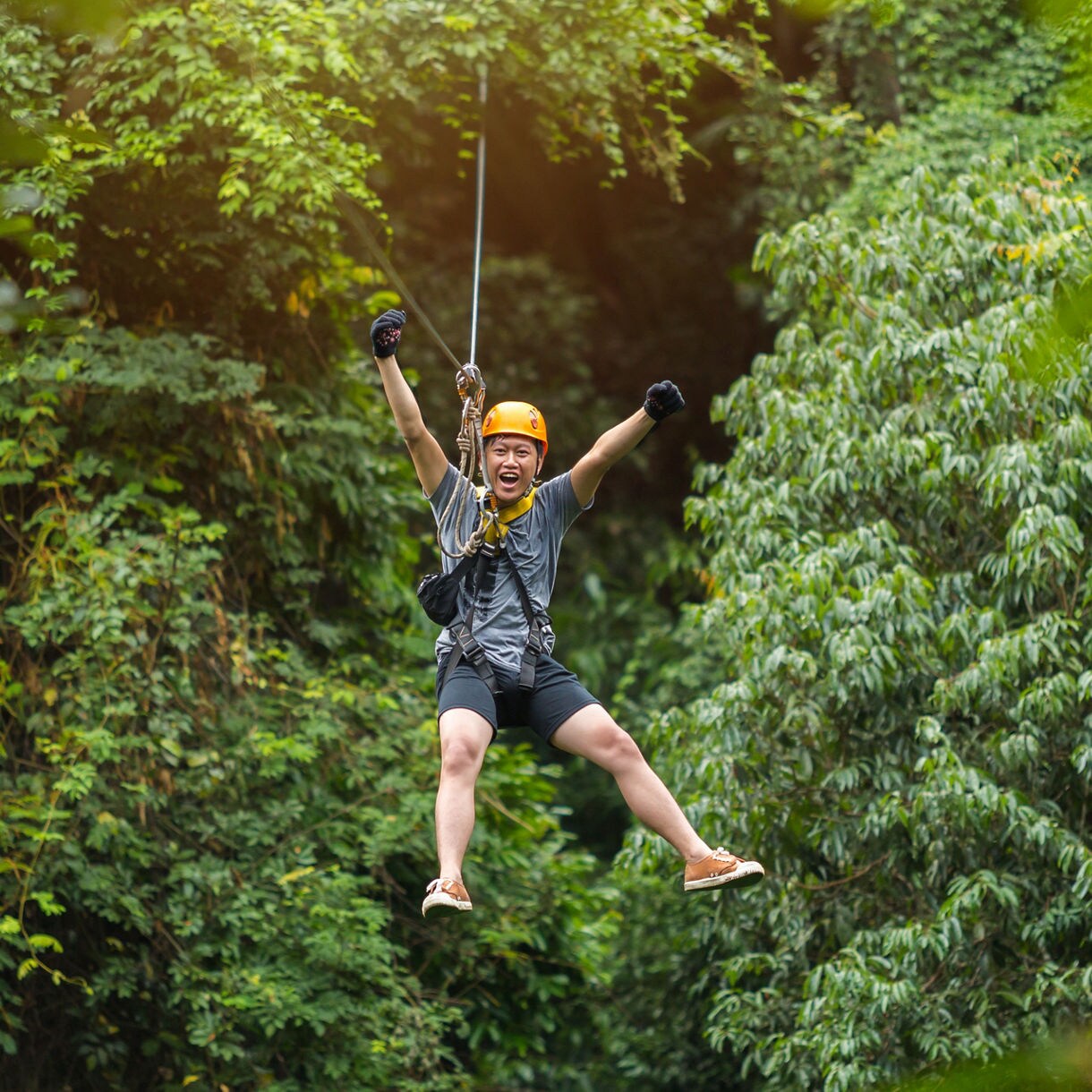 Person ziplining through dense green rainforest in Antigua, wearing a helmet and harness with arms raised in excitement.