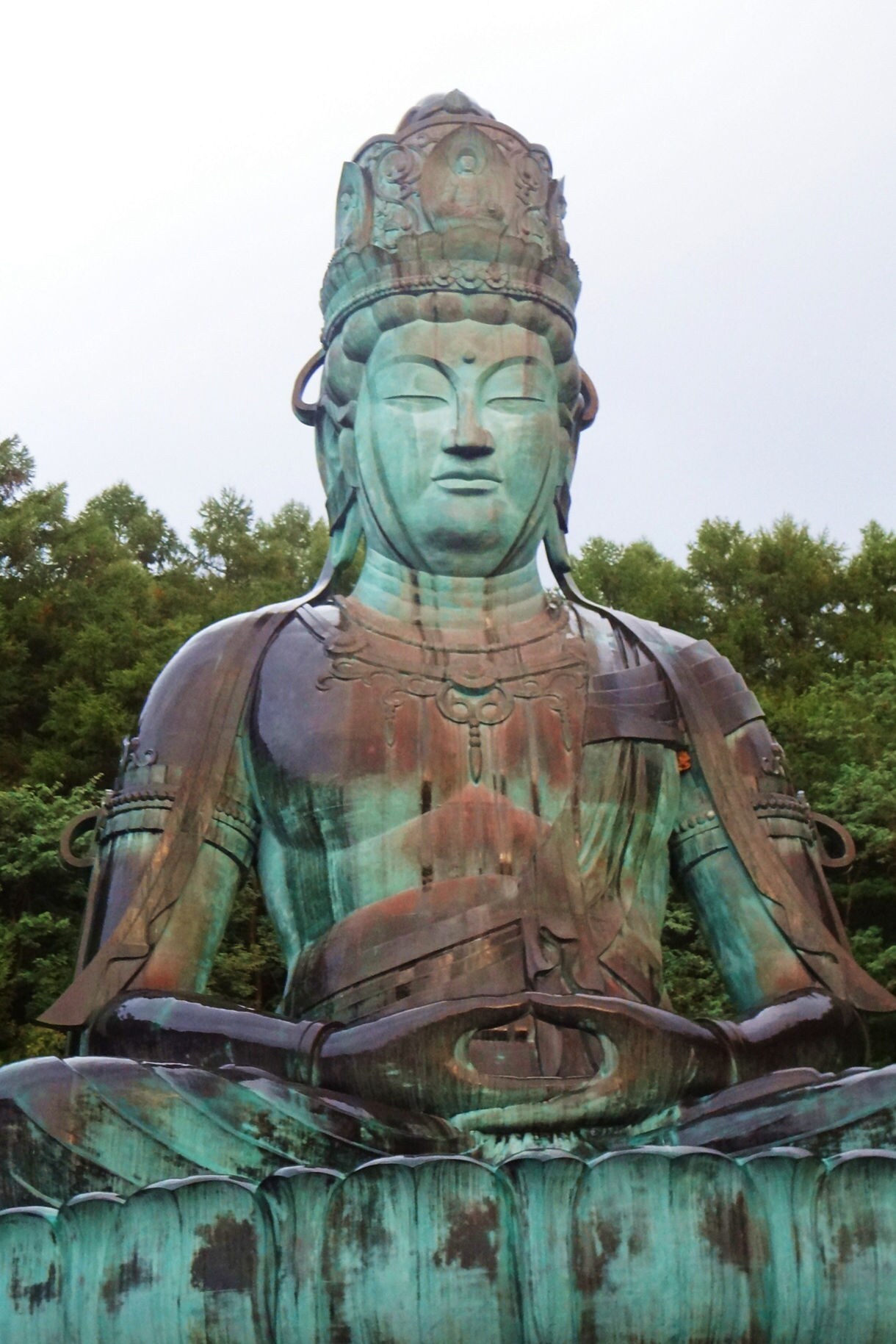 Massive seated bronze Buddha statue with a lotus base, set against tall green trees.