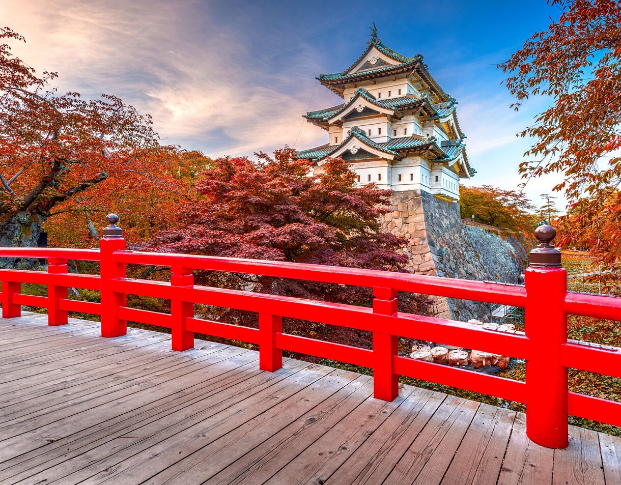 Traditional Japanese castle with green roofs behind red bridge, surrounded by vivid red-orange autumn foliage.