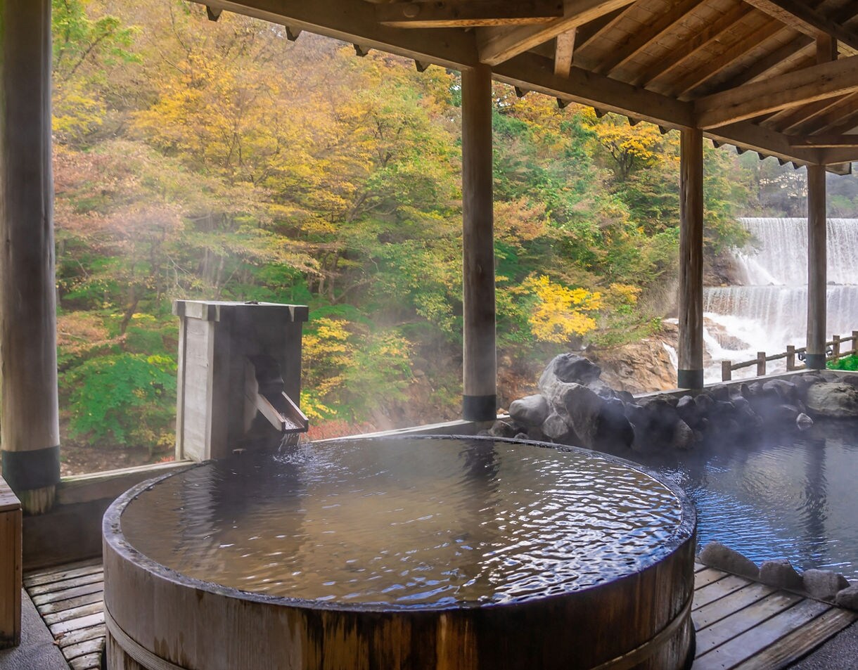 Outdoor hot spring bath with rising steam, wooden roof and view of a forest and waterfall in autumn.