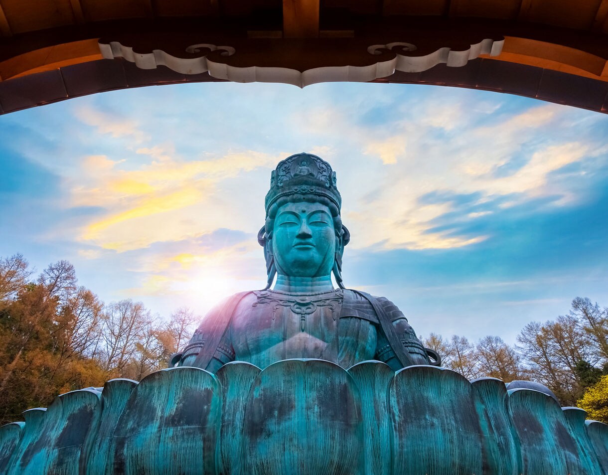 Massive bronze Buddha statue framed by a wooden arch, backlit by sunset, surrounded by trees.