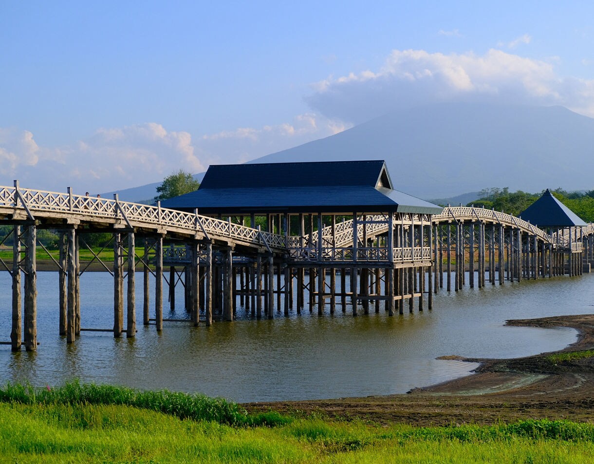 Long arched wooden bridge with covered sections spanning a lake, set against Mt. Iwaki under a hazy blue sky.