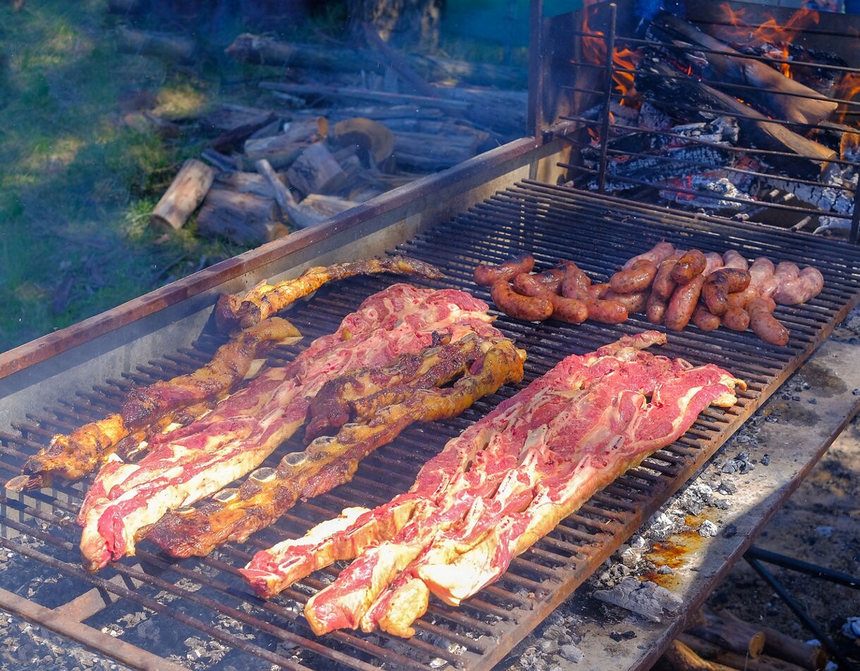 Close-up of an outdoor grill packed with ribs and sausages cooking over wood fire, with smoke rising and logs burning in the background.